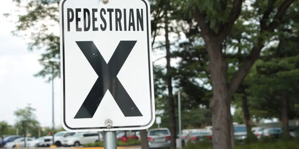 pedestrian x vertical rectangle white black sign on metal post with parking lot trees behind, close up