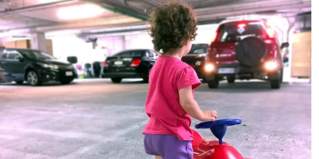 AUCKLAND - FEB 10 2016:Young girl (Naomi Ben-Ari age 2) playing with a toy car in a parking lot while car reversing. In the U.S. at least 50 children are being backed over by vehicles EVERY week.